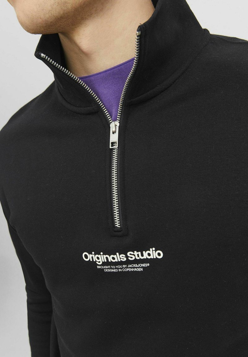 Zwarte quarter-zip sweatshirt van zachte stof met een zilveren rits, witte tekst "Originals Studio" en een paarse onderhemd zichtbaar eronder.