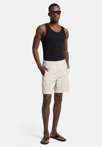 Zwarte tanktop, crèmekleurige shorts met elastische tailleband, slim fit, twee zakken en zwarte teenslippers. Model draagt een zonnebril.