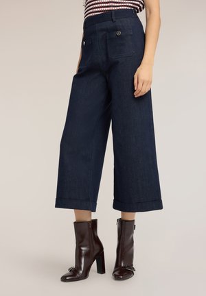 Donna che indossa pantaloni corti a gamba larga di colore scuro con orli risvoltati e stivaletti marrone scuro con tacco, in piedi su uno sfondo semplice.