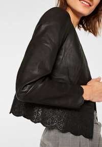 Chaqueta de cuero negro con bajo con volantes y bordados intrincados, mangas largas y una textura suave. No hay cierres visibles.