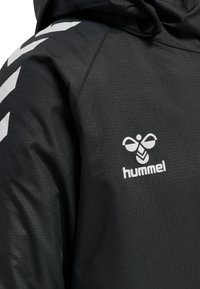 Svart jacka i texturerat tyg, med vitt logo och chevronmönster på axlarna. Inkluderar huva och dragsko.