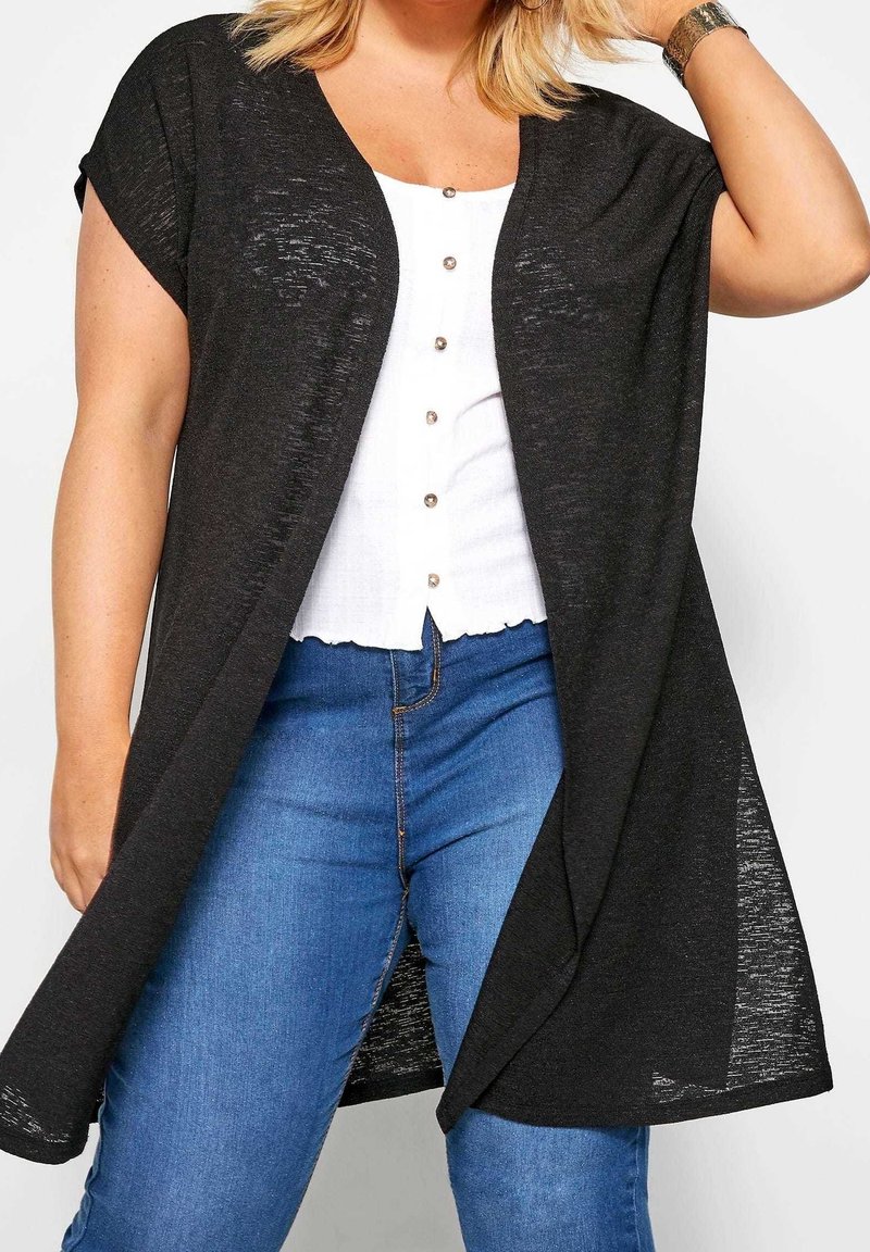 Svart cardigan med öppen front och korta ärmar, tillverkad av lätt material. Har en avslappnad passform. Bärs över en vit skjorta med knappar och blå jeans.