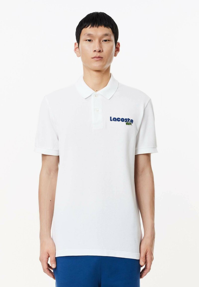 Camisa polo blanca de algodón, con cuello clásico, mangas cortas y logotipo bordado de "Lacoste" con un acento de cocodrilo verde.