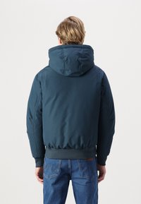 Chaqueta acolchada azul marino con una gran capucha, puños y dobladillo elásticos. Presenta una textura suave y detalles de costura mínimos. Lleva puesta con denim.
