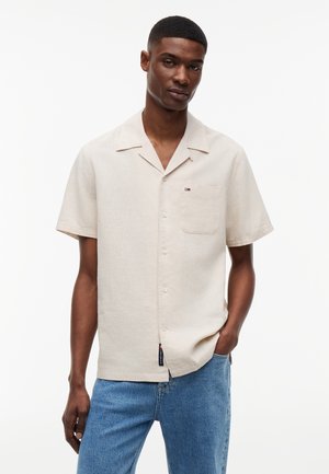 Man die een beige kortärmelig overhemd met knopen en een blauwe spijkerbroek draagt, staand met één hand in de zak tegen een effen witte achtergrond.