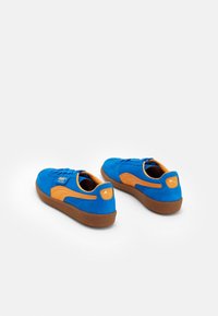 Blå mockaskor med orange accenter, rund tå, snörning och en brun gummisula. Puma-logotyp på sidan.