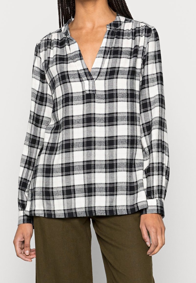Blouse à carreaux noir et blanc avec un col en V peu profond, des manches longues et une coupe décontractée. Confectionnée dans un tissu léger, présentant des motifs à carreaux.