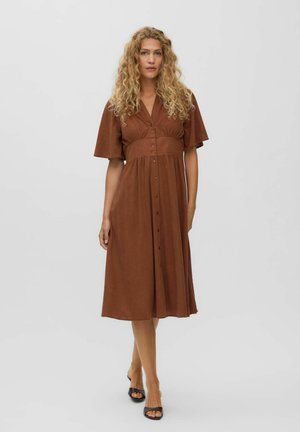 Blonde woman with curly hair wearing a brown buttoned midi dress and black open-toe heels standing against a plain light background.