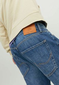 Blå denimjeans med en läderlapp på baksidan av midjan, synlig sömnad och två bakfickor, en med en unik form.