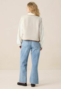 Gilet en polaire beige avec des ouvertures pour les bras, couture contrastée, porté sur une blouse blanche et un jean évasé bleu clair ; vu de dos.