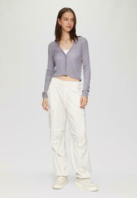 Lichtpaarse geribde cropped cardigan met lange mouwen, over een witte tanktop, gecombineerd met losse witte cargobroek en beige sneakers.