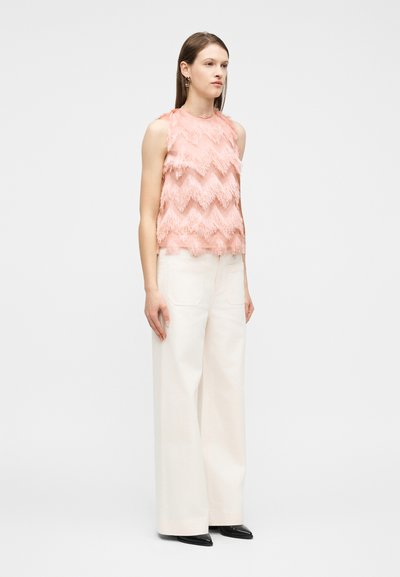 Γυναίκα που φοράει ροζ αμάνικο τοπ με υφή και σχέδιο ζιγκ-ζαγκ, λευκό παντελόνι με φαρδιά μπατζάκια, μαύρα μυτερά παπούτσια και χρυσούς κρίκους στα αυτιά, στέκεται ακίνητη.