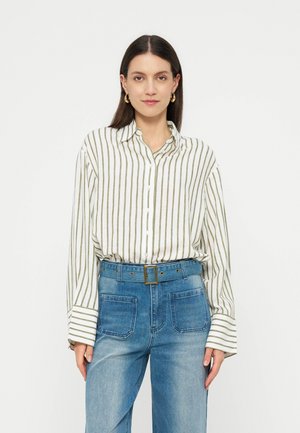 Kobieta o długich ciemnych włosach, ubrana w białą koszulę na guziki z zielonymi pionowymi paskami oraz wysokowaistowe niebieskie dżinsy z paskiem.