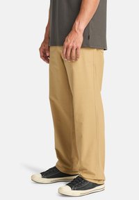 Pantalones de algodón beige con corte recto, que cuentan con bolsillos laterales y un acabado limpio. Combinados con zapatillas negras y una camiseta gris.