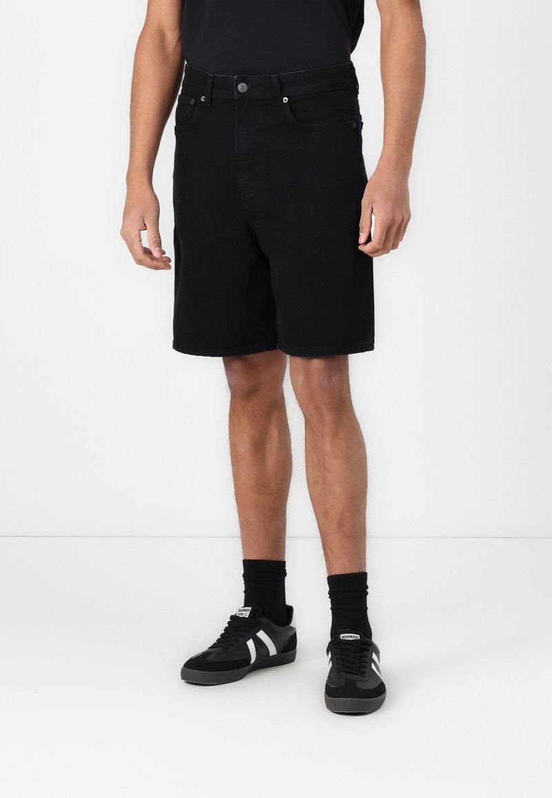 Sorte denimshorts med rett passform, fem lommer og knappestenging, kombinert med svarte sneakers og sokker. Enkelt design.