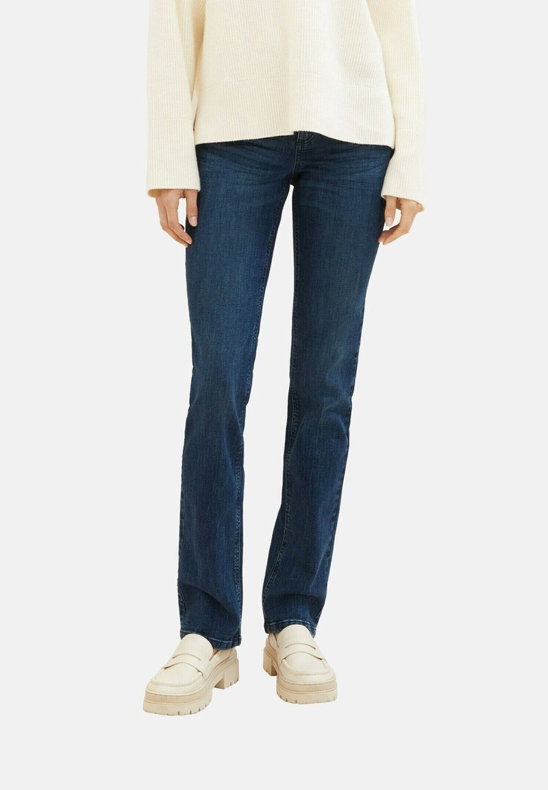 Personne portant un jean bleu foncé droit, un pull crème et des mocassins épais beige clair, debout devant un fond blanc uni.