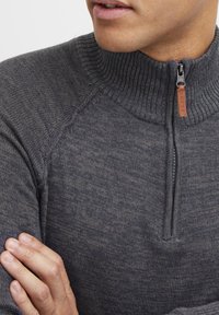Dunkelgrauer Pullover aus Strick mit Viertel-Reißverschluss am Ausschnitt, geripptem Kragen und glatter Textur. Verfügt über Raglanärmel und dezente Musterdetails.