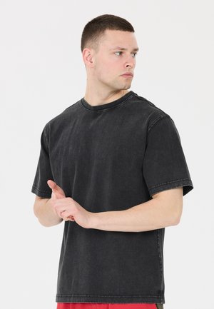 Jonge man in een zwarte vergeelde T-shirt kijkt naar de zijkant met de handen gevouwen voor zich tegen een neutrale achtergrond.