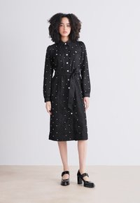Vestito nero con bottoni e cintura in vita, caratterizzato da pois bianchi e un tessuto texturizzato, indossato con scarpe nere lucide con tacco blocco.