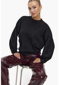 Sweat-shirt noir oversized avec un col montant, associé à un pantalon bordeaux brillant. Le modèle est assis sur une chaise, montrant une bague.