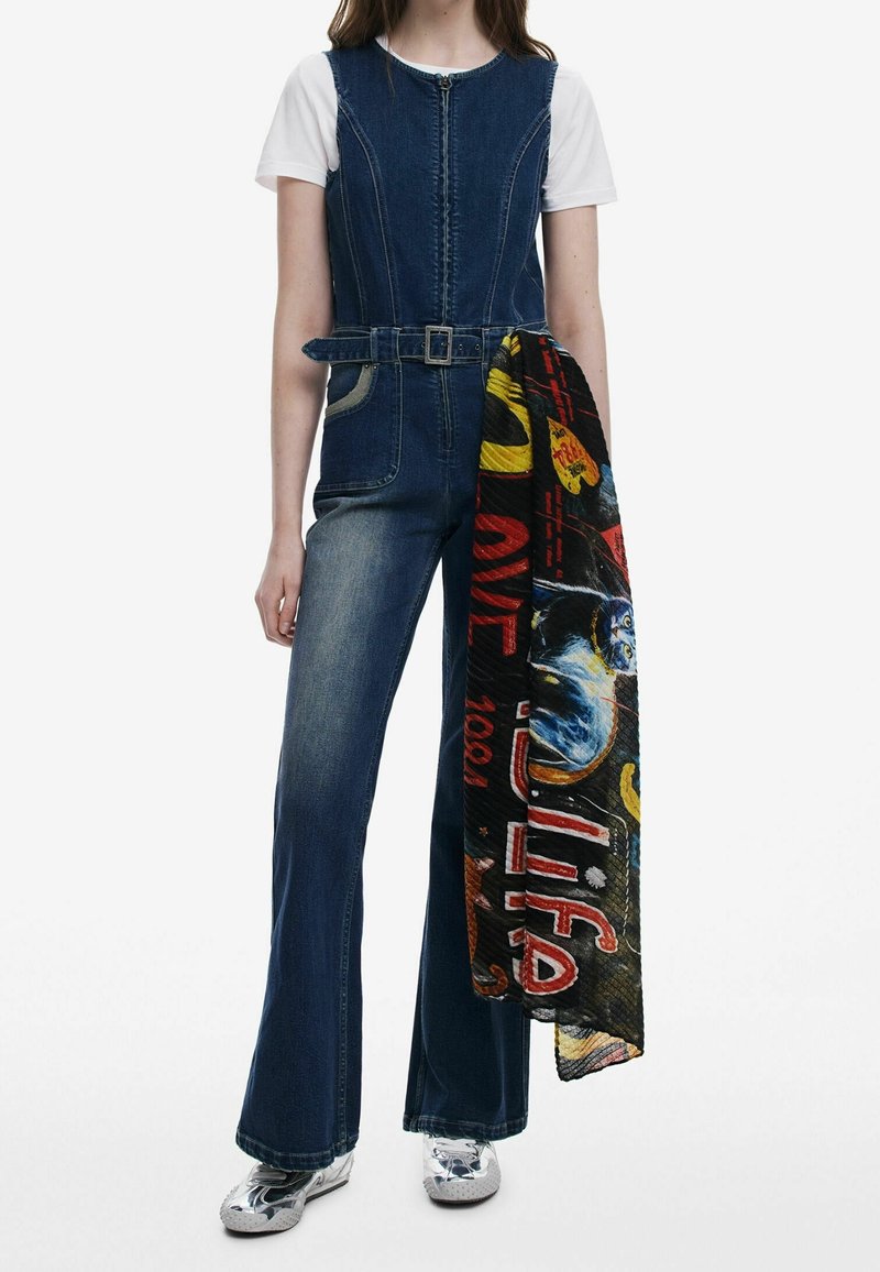 Femme portant une combinaison en jean bleu avec une ceinture, un t-shirt blanc et des chaussures argentées brillantes, tenant une écharpe graphique colorée.