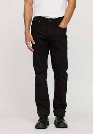 Homme portant un jean droit noir retroussé, un t-shirt blanc, des chaussures noires à lacets et une bague en argent à la main gauche.