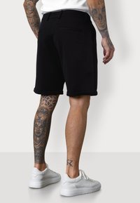 Black cotton shorts with a rolled hem, featuring back pockets and a subtle brand logo. Model is wearing white sneakers.