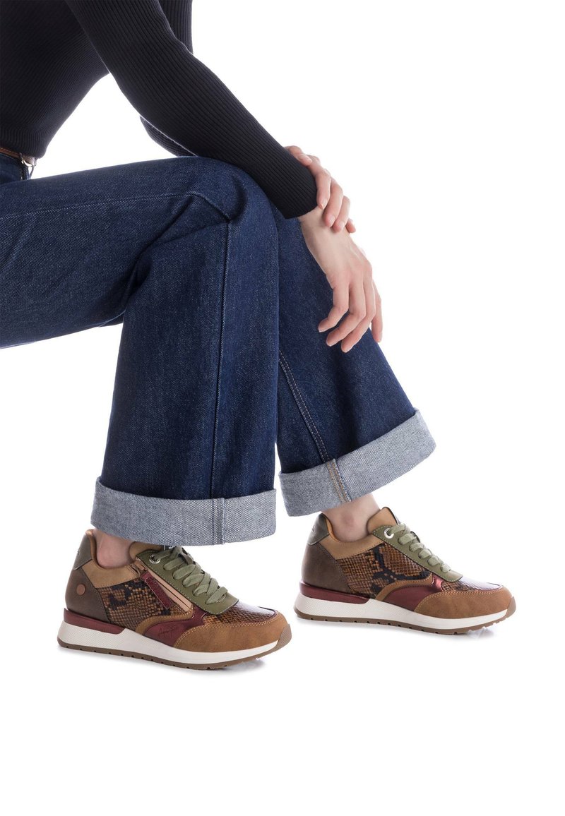 Zapatillas multicolores con cuero marrón y textil verde oliva, que presentan un diseño estampado y acentos en contraste, sobre unos pantalones vaqueros oscuros con puños.