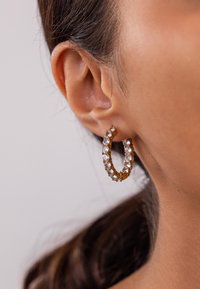 Goldene Hohlschmuck-Ohrringe mit klaren dekorativen Kristallen, die eine gebogene Form und eine strukturierte Oberfläche aufweisen, gesichert mit Metallverschlüssen.