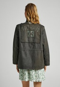 Tmavě zelená džínová bunda s velkou nášivkou na zadní straně, která zobrazuje "Pepe Jeans 73 London." Volný střih, viditelné švy a minimální detaily z kovu.