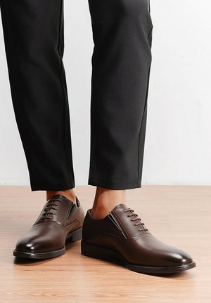 Paire de chaussures habillées en cuir brun foncé poli, portées avec un pantalon noir, debout sur un sol en bois clair.