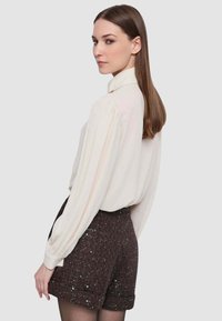 Camicia bianca a maniche lunghe, con una sottile texture e colletto abbottonato. Abbinata a pantaloncini marroni con paillettes, tessuto strutturato e orli risvoltati.