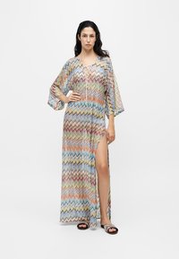 Femme debout portant une longue robe multicolore à motif zigzag avec une fente à l’avant et des sandales à bout ouvert sur fond blanc.