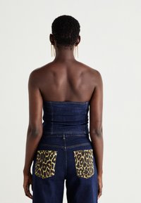 Tamsiai mėlynas be rankovių viršus, turintis prigludusią siluetą, derinamas su tamsiomis džinsomis, turinčiomis geltonus leopardų rašto kišenes. Lygus paviršius, minimalus siūlai.