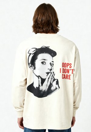 Persona con capelli castano medi indossa una maglietta a maniche lunghe color crema con una donna retrò in bianco e nero con le mani vicino alla bocca e testo rosso "OOPS I DON'T CARE".