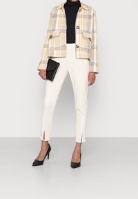 Geruite jas met gele en grijze tinten, zwarte coltrui, crèmekleurige broek met zijsplitten en zwarte hoge hakken, terwijl ze een zwarte clutch vasthoudt.