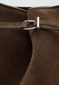 Material de ante marrón con una superficie texturizada, que presenta un detalle de broche metálico plateado en la parte superior, que sujeta el tejido de forma segura.