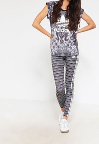 Kvinna som bär en svart och vit Adidas t-shirt med påfågelmönster och matchande leggings med mönster och vita ränder, vita sneakers, händerna på höfterna.