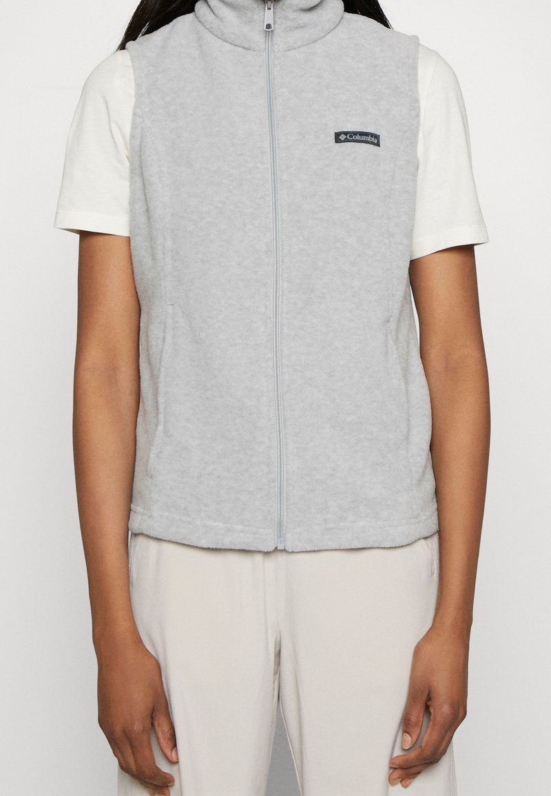 Persoon in een lichtgrijze ritsvest van fleece over een wit T-shirt met korte mouwen en lichtgekleurde broek, staande tegen een eenvoudige achtergrond.