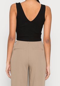 Schwarzes geripptes Tanktop mit einem V-förmigen Rückendetail, kombiniert mit beigen hochgeschnittenen Hosen, die über Gesäßtaschen und eine glatte Textur verfügen.