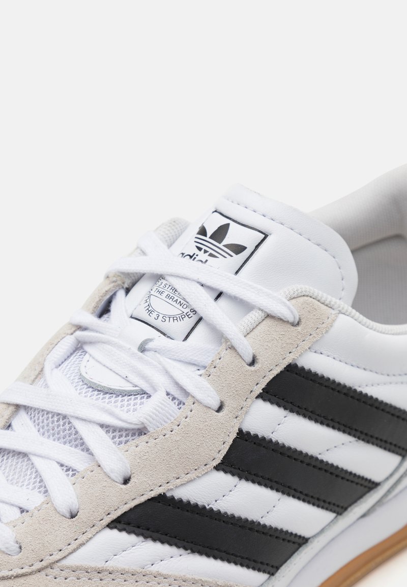 Weißer Sneaker aus Leder und Wildleder mit schwarzen Streifen und weißen Schnürsenkeln. Auffälliges Logo auf der Zunge und strukturiertes Sohlenmuster.