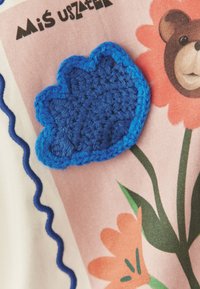 Niebieska kwiatowa łatka na tkaninie wykonana szydełkiem, przedstawiająca wzory kwiatowe i misie, z różowymi i zielonymi akcentami. Dekoracyjna falista niebieska lamówka wzdłuż krawędzi.