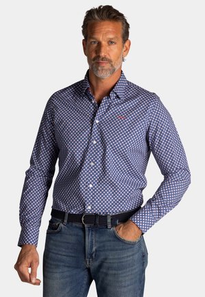 Man met kort haar en baard, gekleed in een blauw patroon overhemd met lange mouwen, ingestopt in een blauwe spijkerbroek met een zwarte riem, hand in de zak.