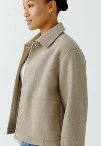 Beige Wollmischung Jacke mit Kragen, Druckknöpfen, langen Ärmeln und einer Seitentasche, vor einem hellen Hintergrund präsentiert.