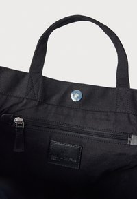 Μαύρη τσάντα tote με υφασμάτινο σώμα, επάνω χερούλια, κούμπωμα με πατητήρα και εσωτερική τσέπη με φερμουάρ. Διαθέτει μικρή μαύρη δερμάτινη ετικέτα.