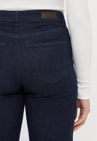 Tummat farkut korkealla vyötäröllä, jossa on ruskea nahkainen brändilogo ja kaksi takataskua hienovaraisella tikkausyksityiskohdalla.