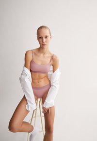 Soutien-gorge rose clair et culotte assortie avec une marque discrète, portés sous une chemise blanche ample, assis sur un tabouret en bois contre un fond pâle.