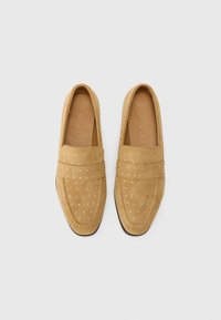 Zamszowe loafery w kolorze tan z ozdobnymi złotymi ćwiekami, zaokrąglony czubek, przeszycia na wierzchu cholewki oraz płaska czarna podeszwa.