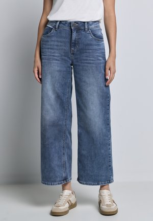 Wijde blauwe jeans van denimstof met een vervaagd effect. Beschikt over vijf zakken en een knoopsluiting. Gecombineerd met lichtgekleurde sneakers.