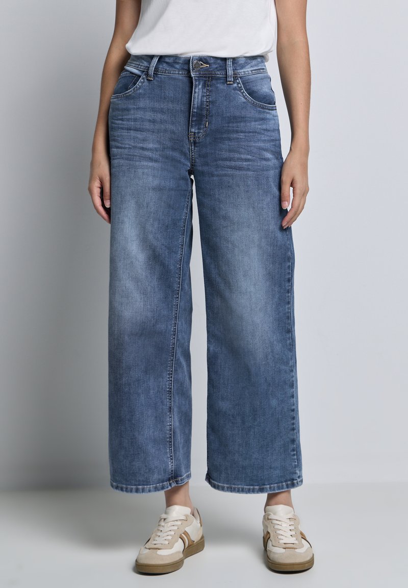 Jeans bleu à jambes larges en tissu denim avec une finition décolorée. Dotés de cinq poches et d'une fermeture à bouton. Associés à des baskets de couleur claire.